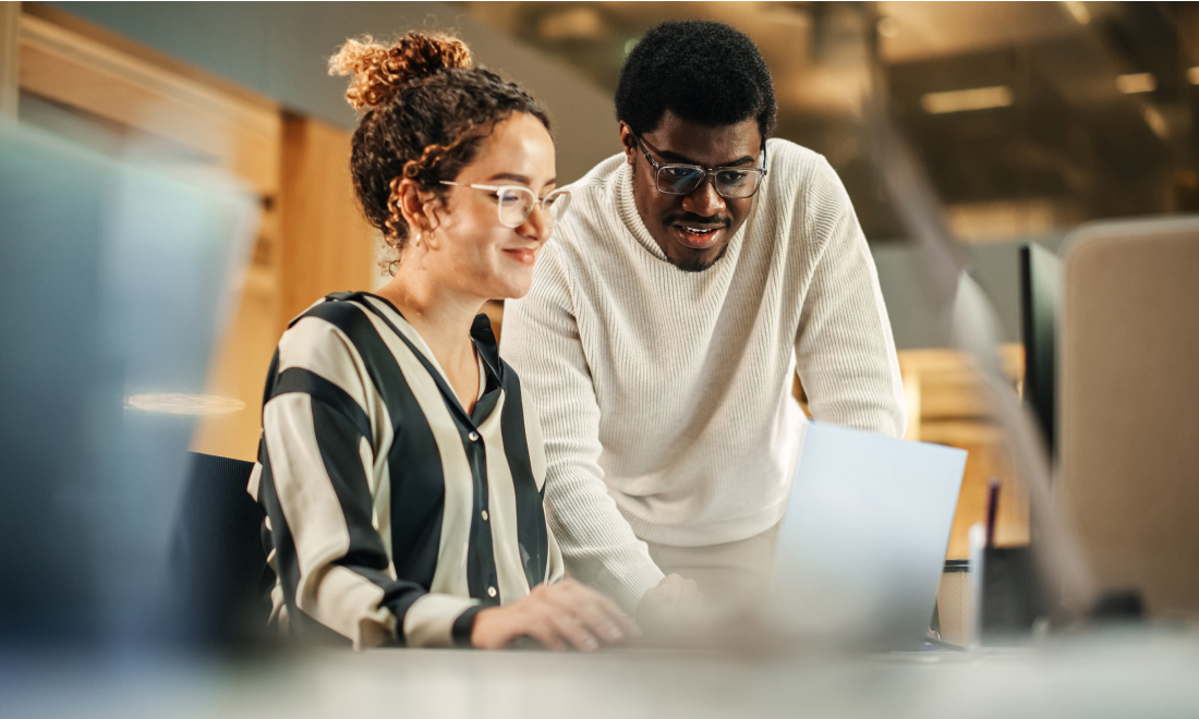 a woman sitting looking at a laptop while a man stands beside her, also looking at the laptop