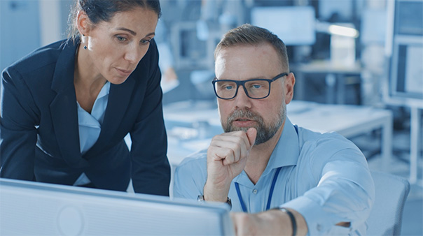 photo of a man and a woman in an office. The man is seated and pointing at a computer screen in the foreground, while the woman is looking at the same screen from over his shoulder.