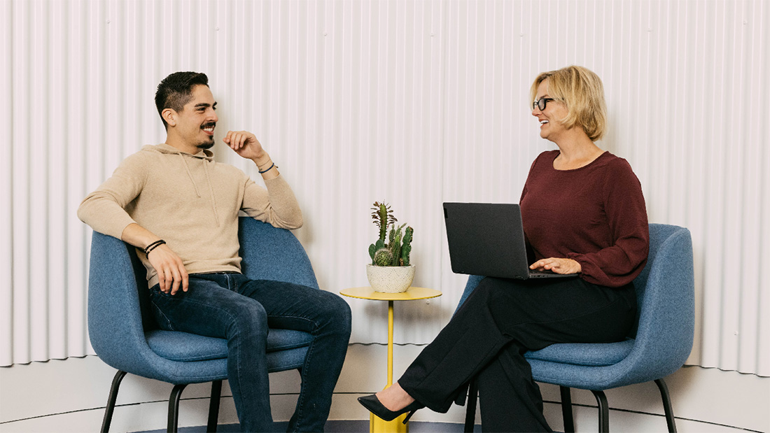 a man and woman sitting on blue padded chairs in front of a white wall. they're looking at each other and smiling, while the woman has a laptop open on her lap.