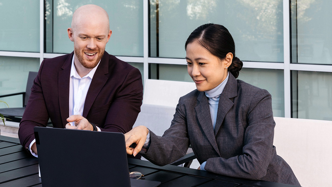 a man and a woman wearing suits, sitting outside in front of a laptop, pointing to something on the screen